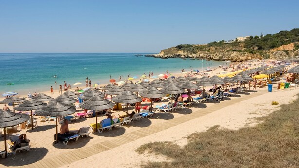 A beach with rows of sun loungers and umbrellas, a wooden walkway, and a rocky coastline in the background. - Spacious 4 bedroom villa with pool, near beach, ideal for families (Olhos de Água)