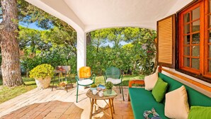 Un patio techado con un sofá verde, sillas de mimbre y una mesa con una cesta de fruta.