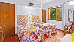 Una habitación con dos camas, una puerta de madera, un ventanal con vistas a la vegetación y una mesita con una lámpara.