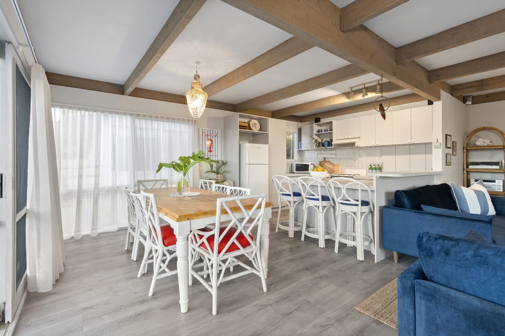A spacious living area with a wooden dining table surrounded by white wicker chairs, a blue sofa, and a kitchen area with white cabinets and a microwave.