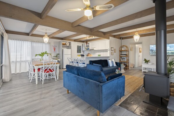 A spacious living area with a blue sofa, dining table, and chairs. There is a kitchen area with white cabinets and a stove. The room has wooden beams on the ceiling and a large window with sheer curtains.