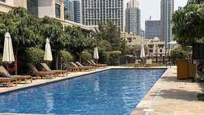 A swimming pool with lounge chairs and umbrellas, surrounded by greenery and tall buildings in the background. - Durrani Homes - Arabian luxury at Souk Al Bahar besides Burj Khalifa & Dubai Mall (Dubai)