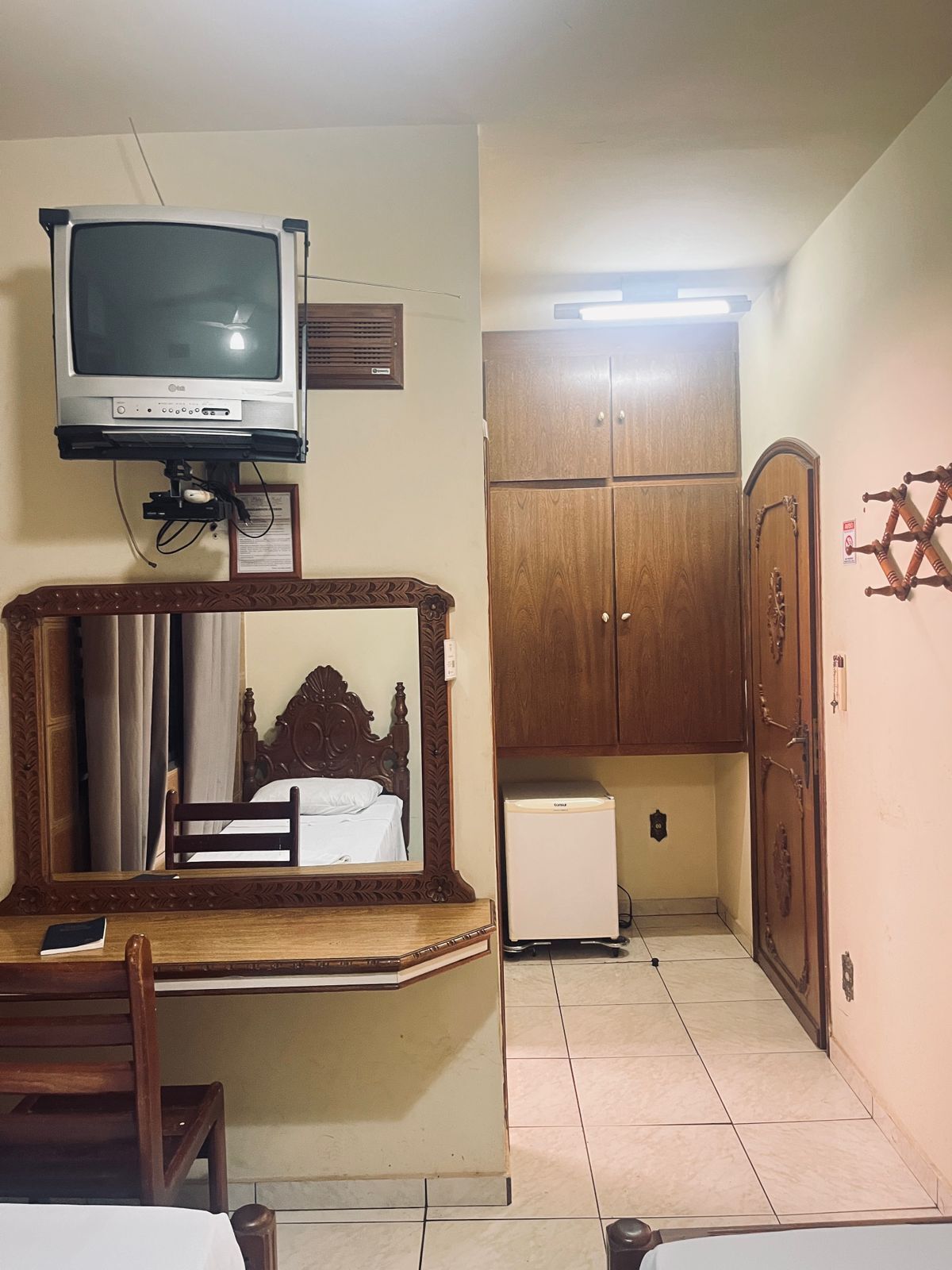 A hotel room with a bed, a wooden cabinet, a small refrigerator, a television mounted on the wall, and a decorative mirror.