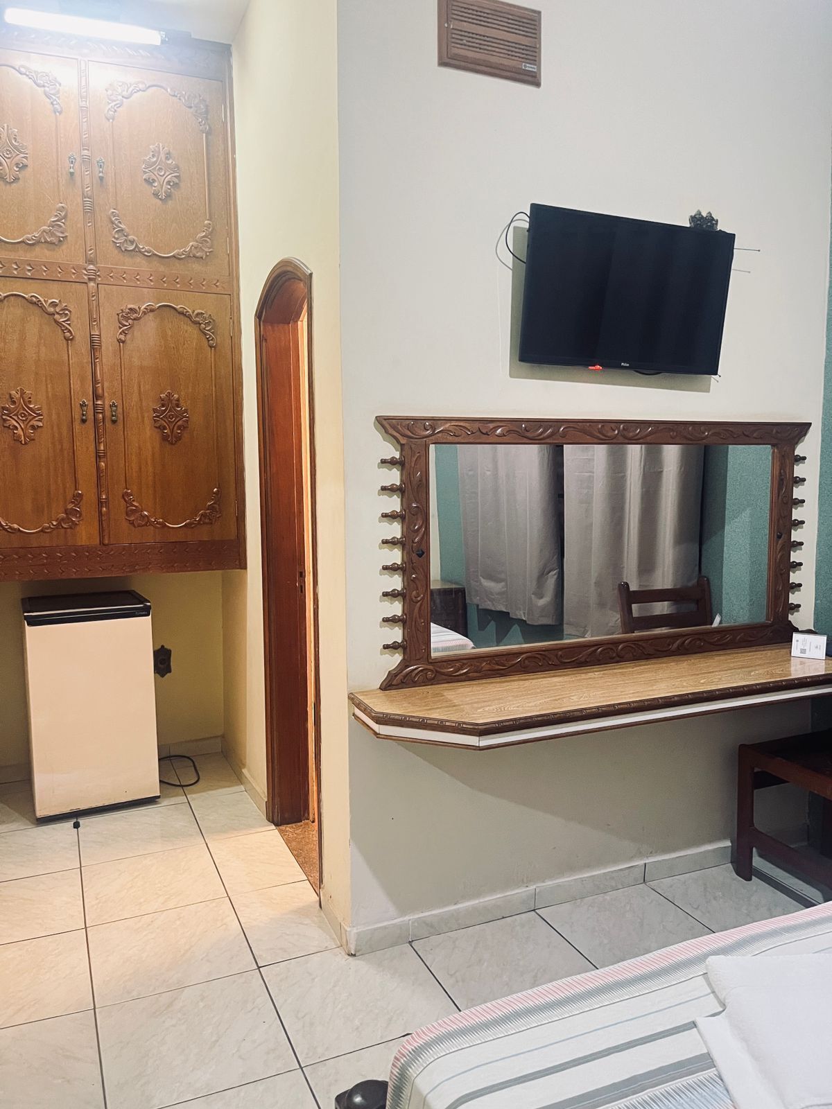A bedroom with a wooden wardrobe, a small refrigerator, a mirror with a wooden frame, and a television mounted on the wall.