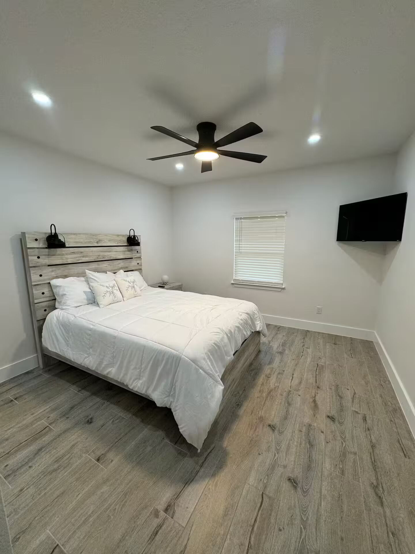A bedroom with a bed, a ceiling fan, a TV mounted on the wall, and a window with blinds.