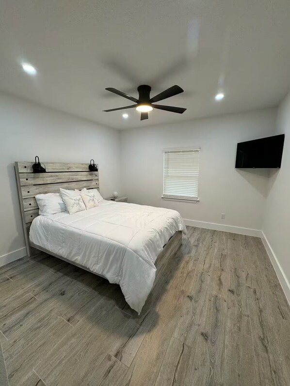 A bedroom with a bed, a ceiling fan, a TV mounted on the wall, and a window with blinds.