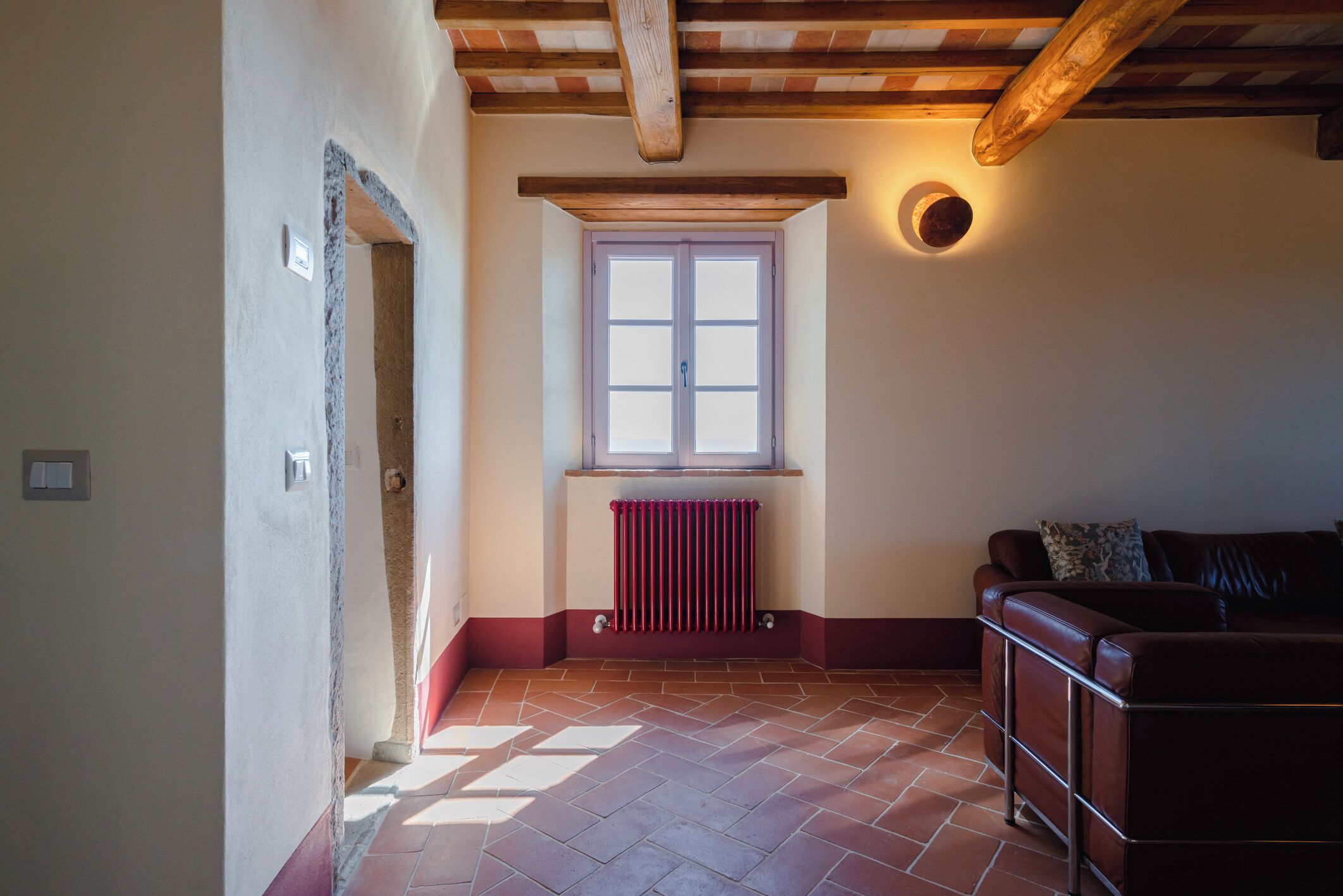 Una habitación con piso de baldosas, una ventana con radiador debajo, una viga de techo de madera y un sofá de cuero marrón.