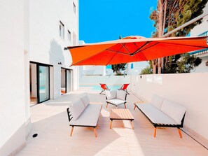 Un espace extérieur moderne, aménagé avec des canapés blancs et une table en bois, un parasol rouge et une piscine en arrière-plan.