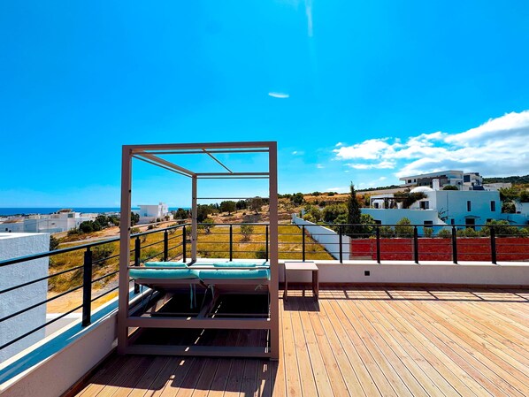 A wooden deck with a metal-framed sunbed and a table, overlooking a coastal landscape with buildings and a clear sky. - Villa Alba Luxe et Confort a Hammamet (Hammamet)