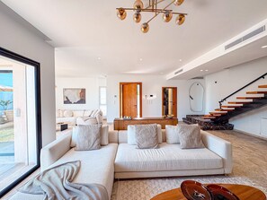 Ein geräumiges Wohnzimmer mit einer großen Eckcouch, einem Couchtisch und einem Essbereich mit Holztisch und Stühlen. Eine Treppe mit Holzstufen und einer schwarzen Geländer führt in eine obere Ebene.