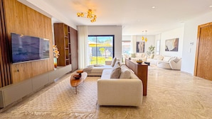 A modern living room with a large flat-screen TV mounted on a wooden wall, a beige sofa, a coffee table, and a wooden cabinet. - Villa Alba Luxe et Confort a Hammamet (Hammamet)