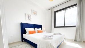 A bedroom with a bed, white bedding, a blue headboard, two framed pictures on the wall, a window with curtains, and a patterned rug. - Petit Cocon Neuf et Styl la Marsa (La Marsa)