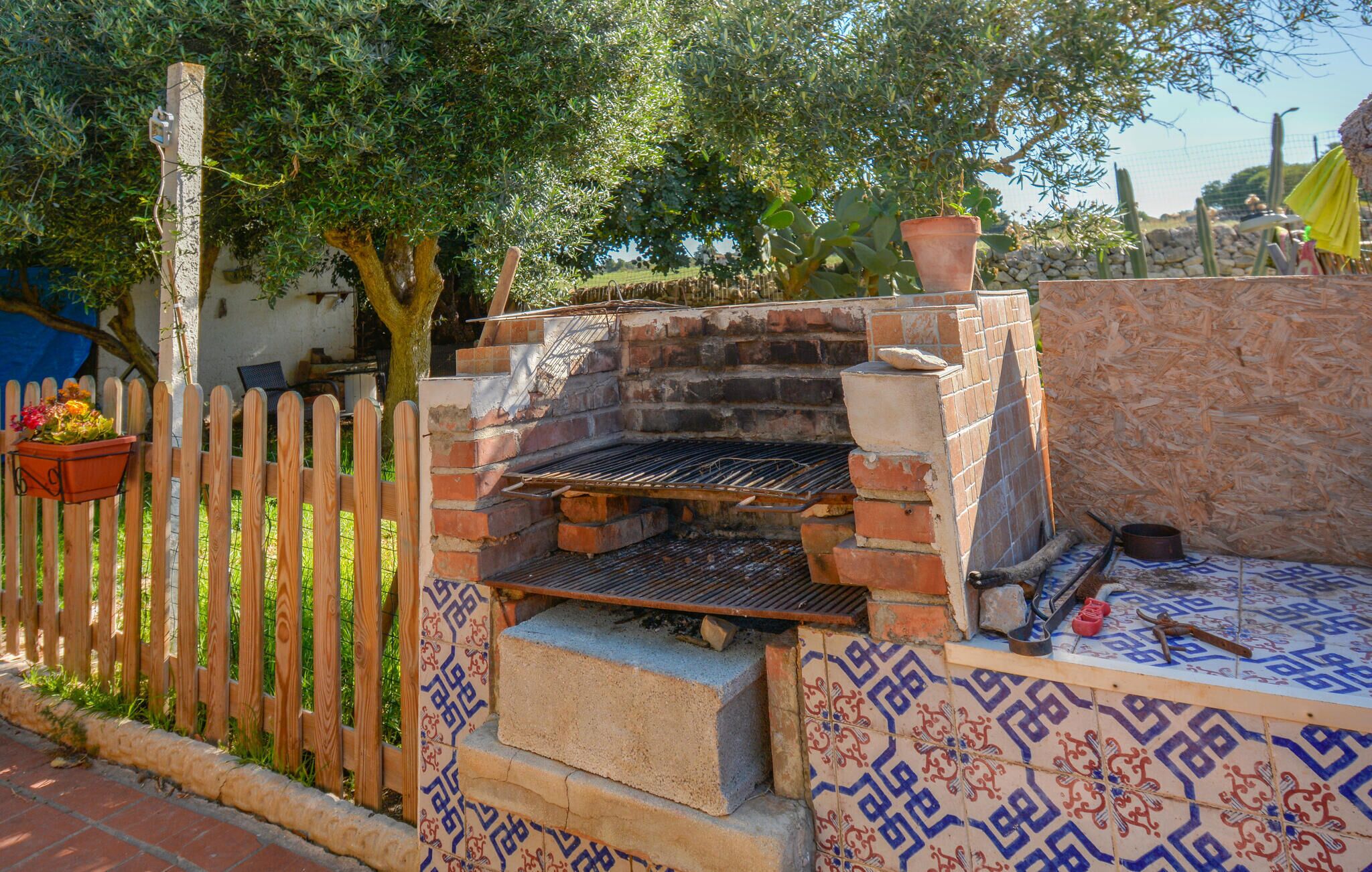 An outdoor brick oven barbecue area with a tiled floor, a wooden fence, and a potted plant.