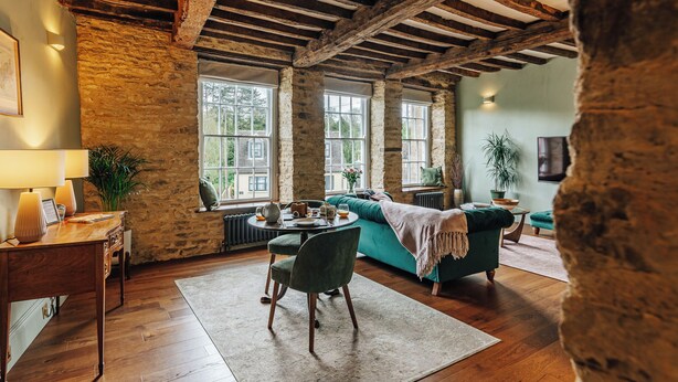 A rustic room with exposed wooden beams, a green sofa, a round table, and a view of the outside through large windows. - NEW luxury historic apartment, private garden, parking, dog-friendly, in Tetbury (Tetbury)