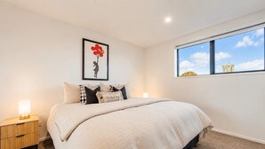 Un dormitorio con una cama grande, una mesita de noche, una obra de arte enmarco y una ventana con vista a la ciudad.