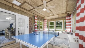 Ett bordtennisbord i ett rum med synliga träbjälkar i taket, stora fönster och randiga gardiner.