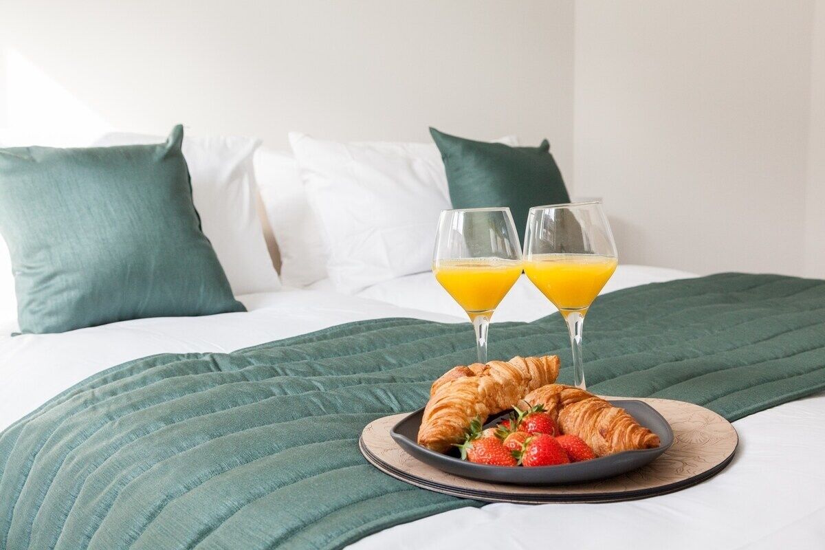 Ein ordentlich bezogenes Bett mit grüner Decke und Kissen, ein Teller mit Croissants und Erdbeeren sowie zwei Gläser Orangensaft.