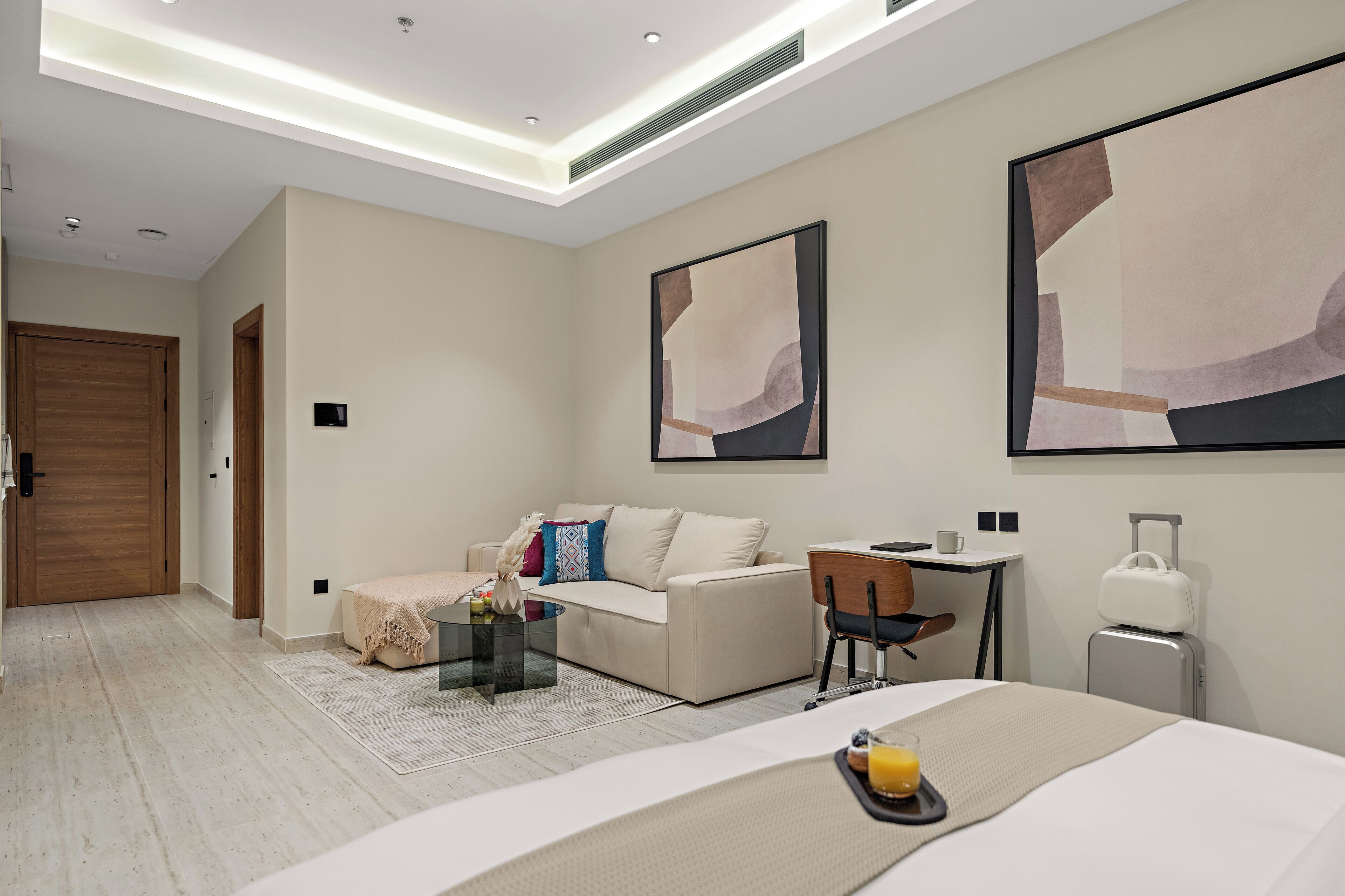 Ein modernes Hotelzimmer mit einem Bett, einer Couch, einem Schreibtisch und einem Stuhl. Ein Koffer und ein kleiner Tisch mit einem Glas Saft sind ebenfalls vorhanden.