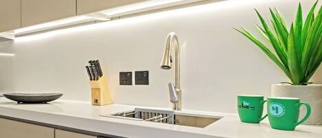 A modern kitchen with a stainless steel sink, a knife block, a potted plant, and two green mugs on the counter.