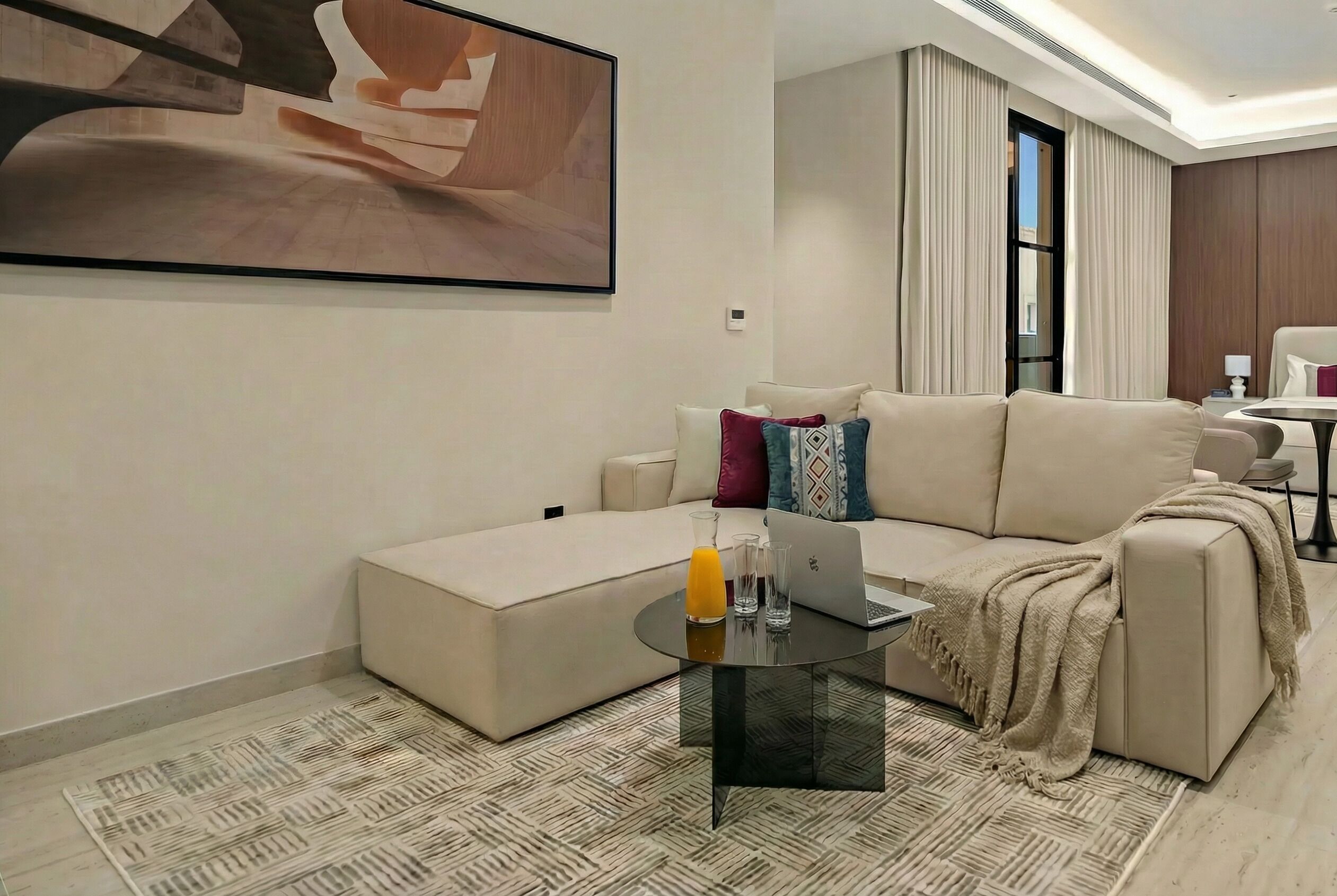 A modern living room with a sofa, a glass coffee table, a laptop, and a painting on the wall.