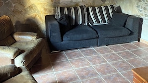 Ein Zimmer mit Steinwänden, einem dunkelgrauen Sofa und gestreiften Kissen, einer gelben Hängeleuchte und einem Fliesenboden.