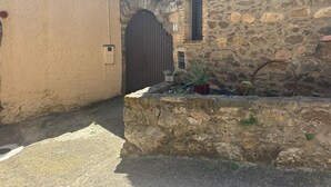 Ein schmaler Gang mit einer Steinmauer auf einer Seite und einer beigen Mauer auf der anderen. Ein großes Holztor und ein kleines Fenster mit einer Pflanze sind in die Steinmauer eingelassen.