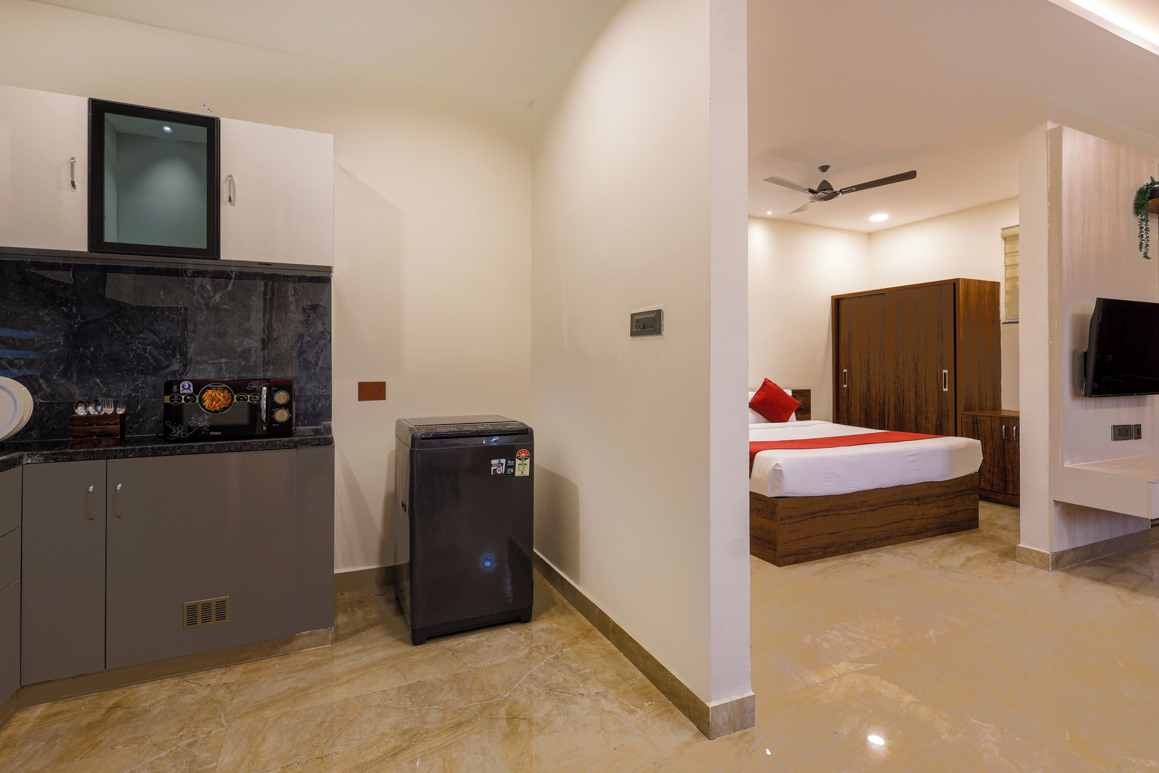 A compact hotel room with a kitchenette, a bed with a red pillow, a TV, and a ceiling fan.