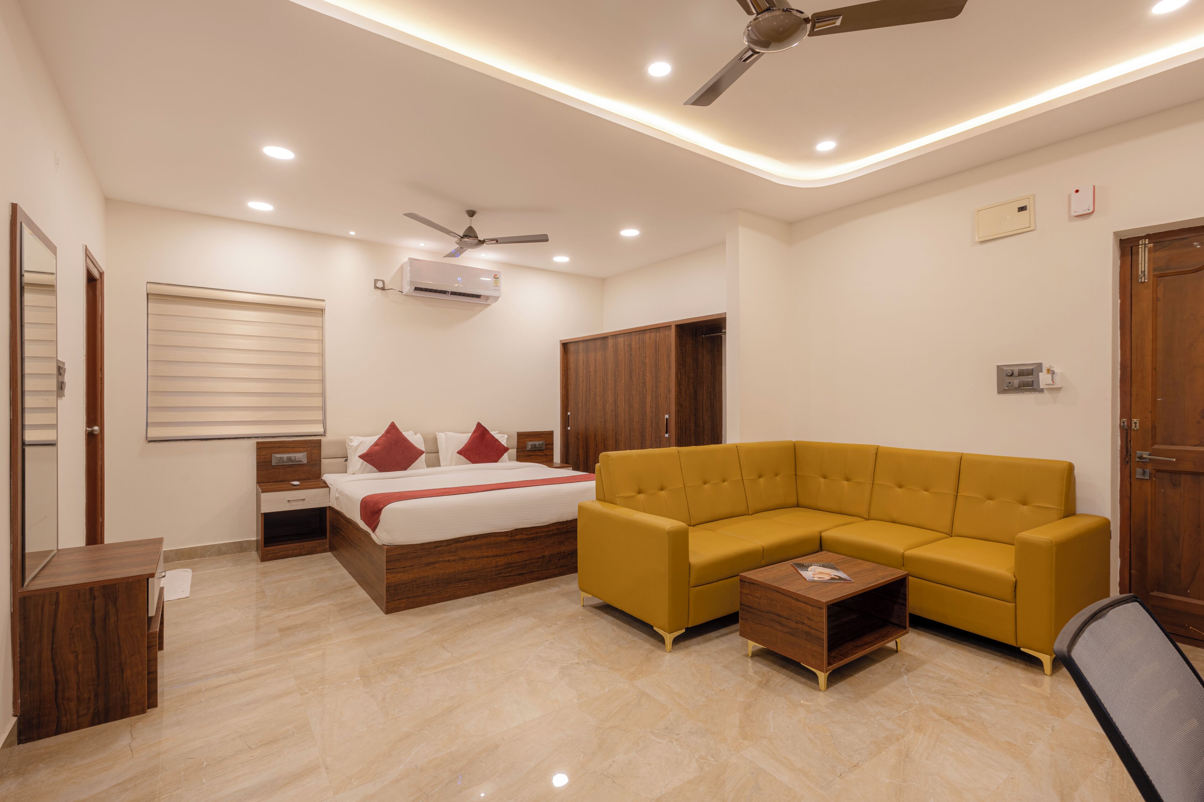 A modern hotel room with a bed, a yellow sofa, a wooden nightstand, a ceiling fan, and recessed lighting.