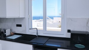 Μια σύγχρονη κουζίνα με μαύρο πάγκο, λευκά ντουλάπια, έναν νεροχύτη και ένα παράθυρο με θέα στη θάλασσα.