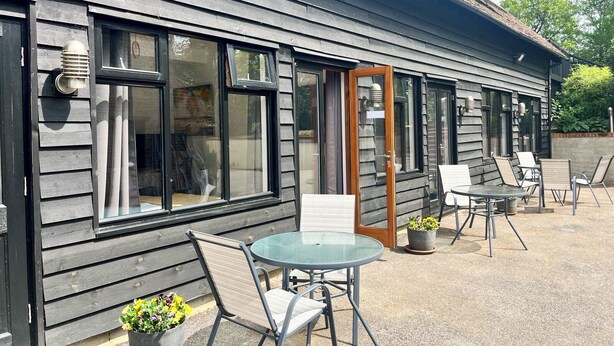 Un bâtiment à l’extérieur en bois foncé, doté de grandes fenêtres et d’une porte en verre donnant sur un espace extérieur aménagé avec des chaises en métal et une table ronde en verre.