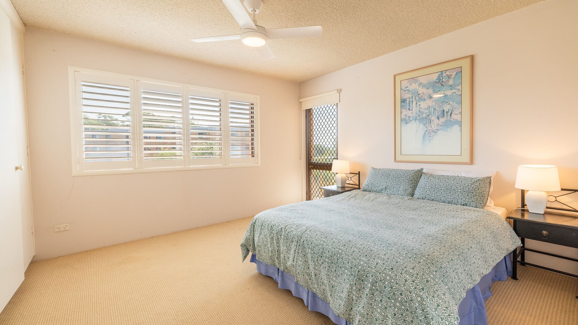Ein Schlafzimmer mit einem Bett, zwei Nachttischen mit Lampen, einem Deckenventilator, einem Fenster mit Jalousien und einem Gemälde an der Wand.