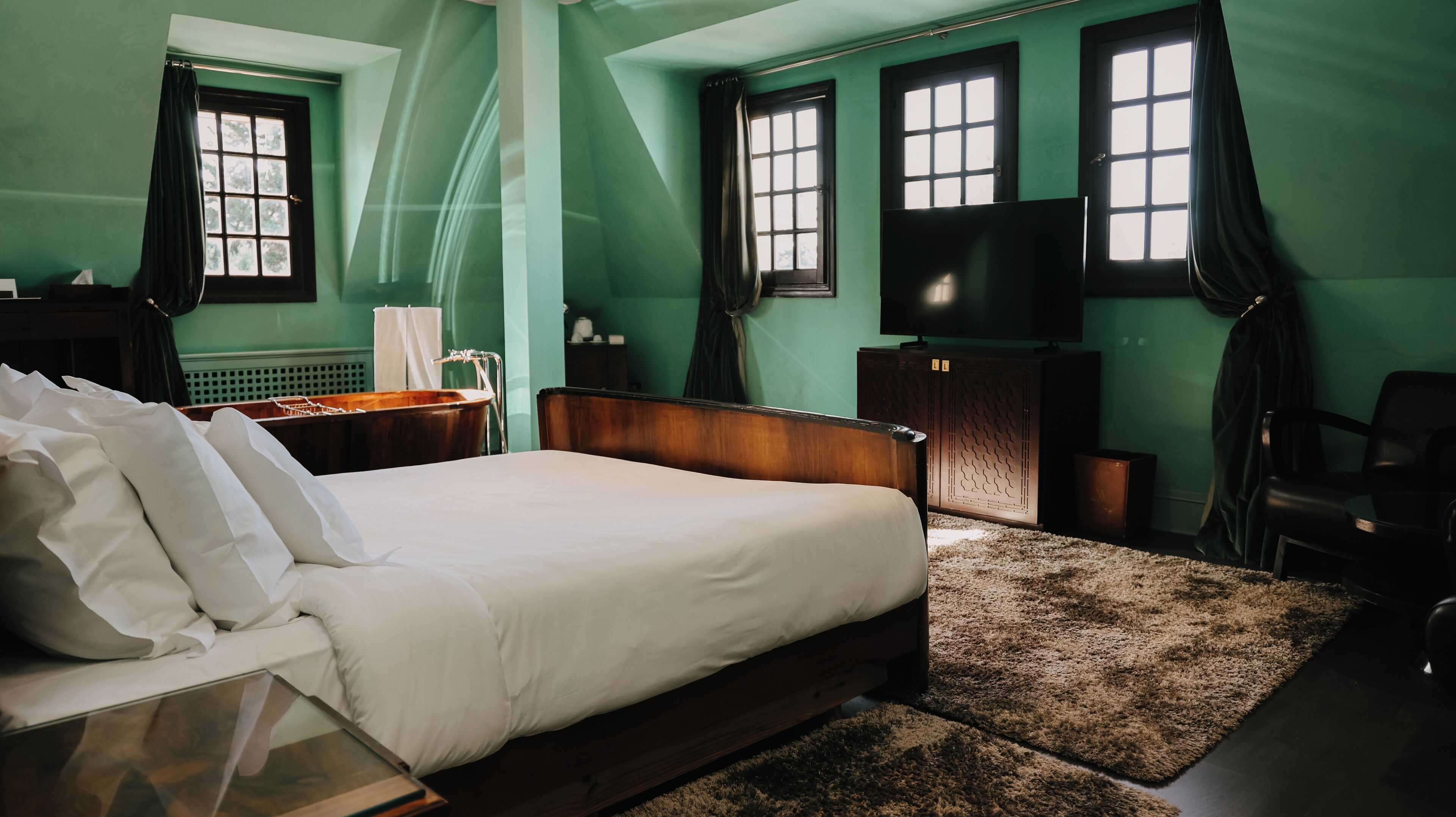 Une chambre à coucher avec un grand lit, une tête de lit en bois, une table de chevet en bois, un bureau, une chaise, un téléviseur et une fenêtre avec des rideaux.