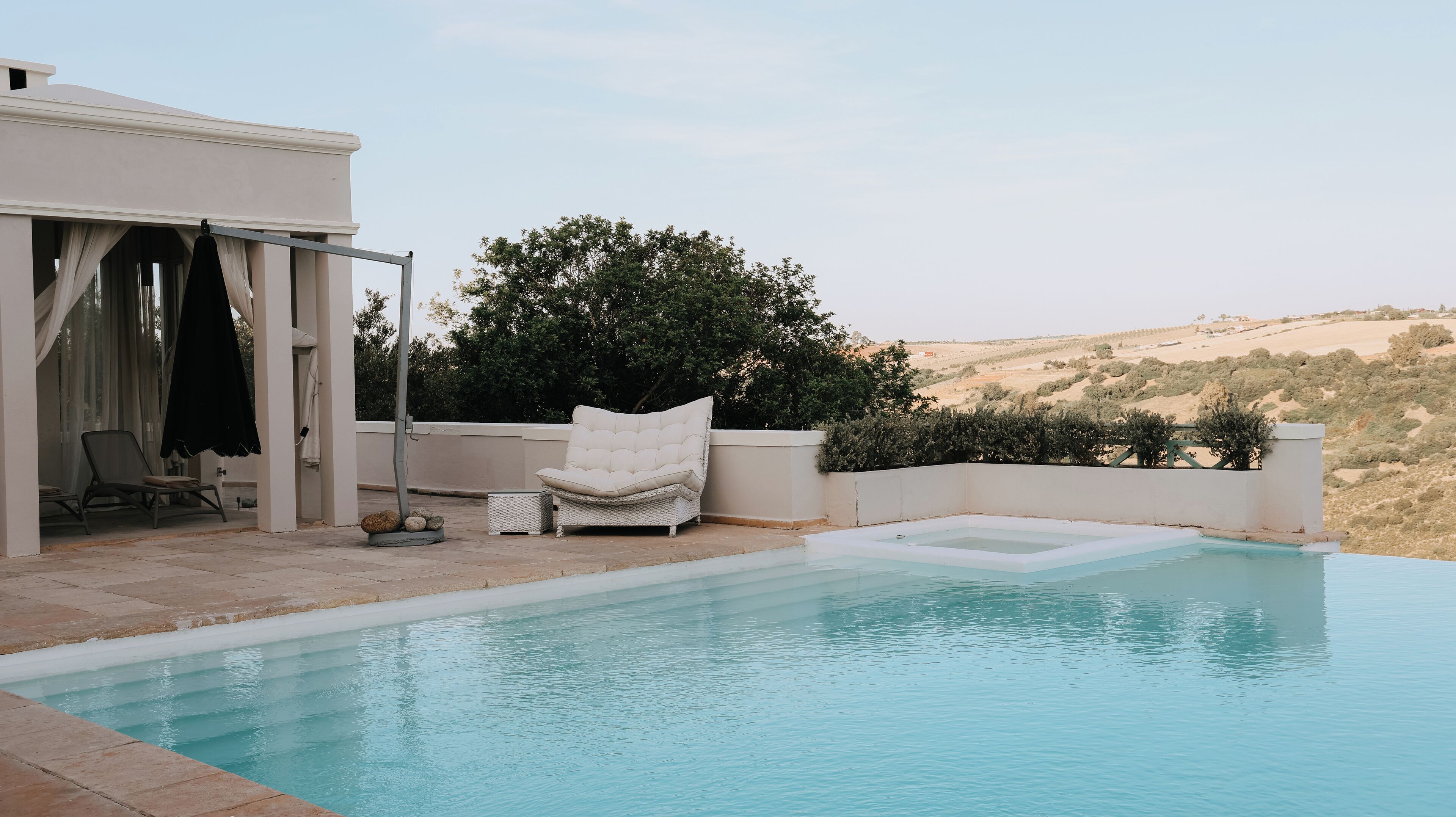 A pool area with a white chair, a covered seating area, and a view of the countryside.
