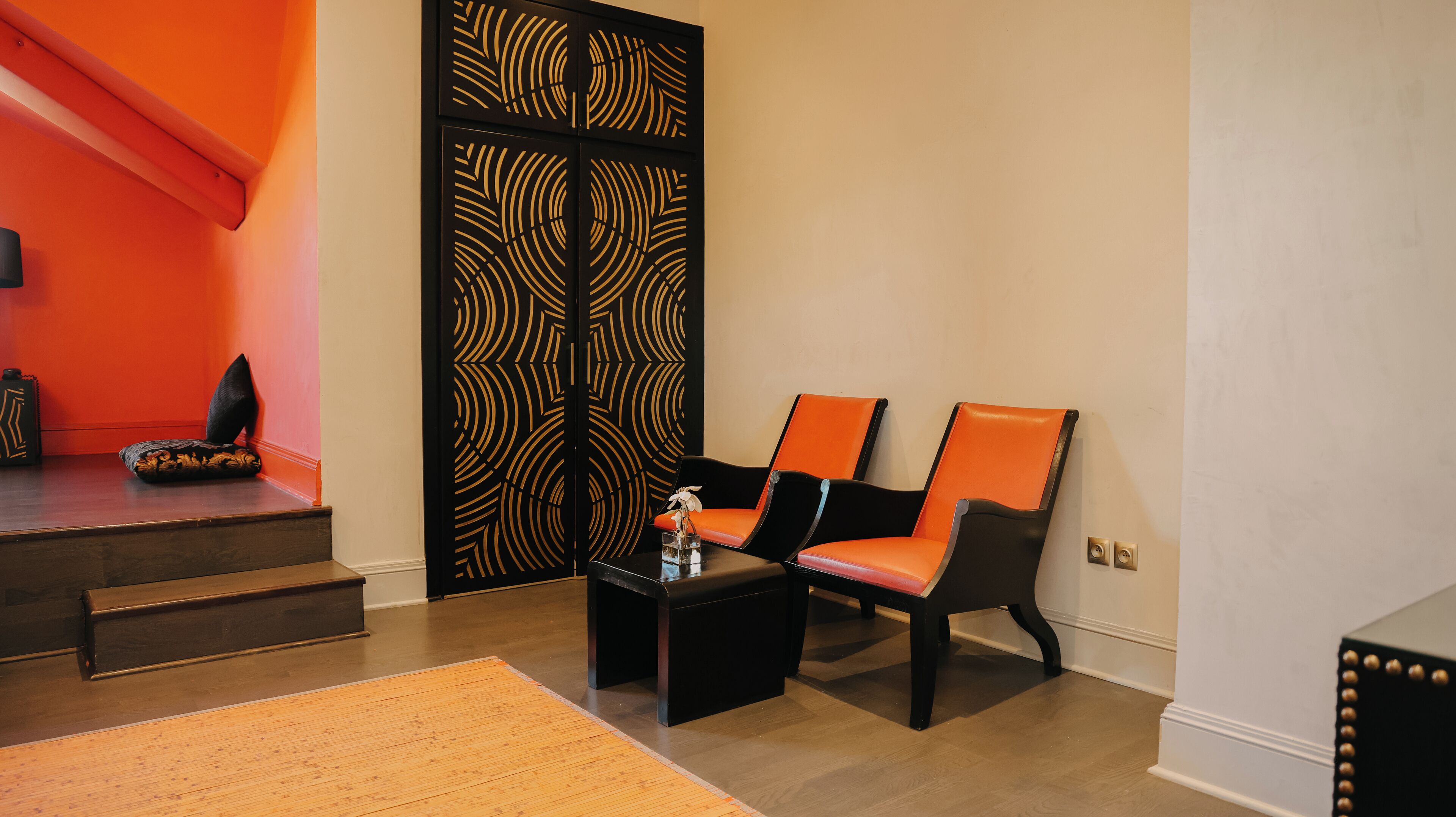 A room with two orange chairs, a black table with a vase, and a patterned black partition.