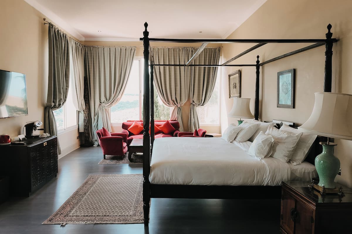 Ein Bett mit Baldachin, ein Nachttisch, eine Lampe und ein Fernseher auf der linken Seite. In der Mitte befindet sich ein rotes Sofa und ein kleiner Tisch mit einer Lampe.
