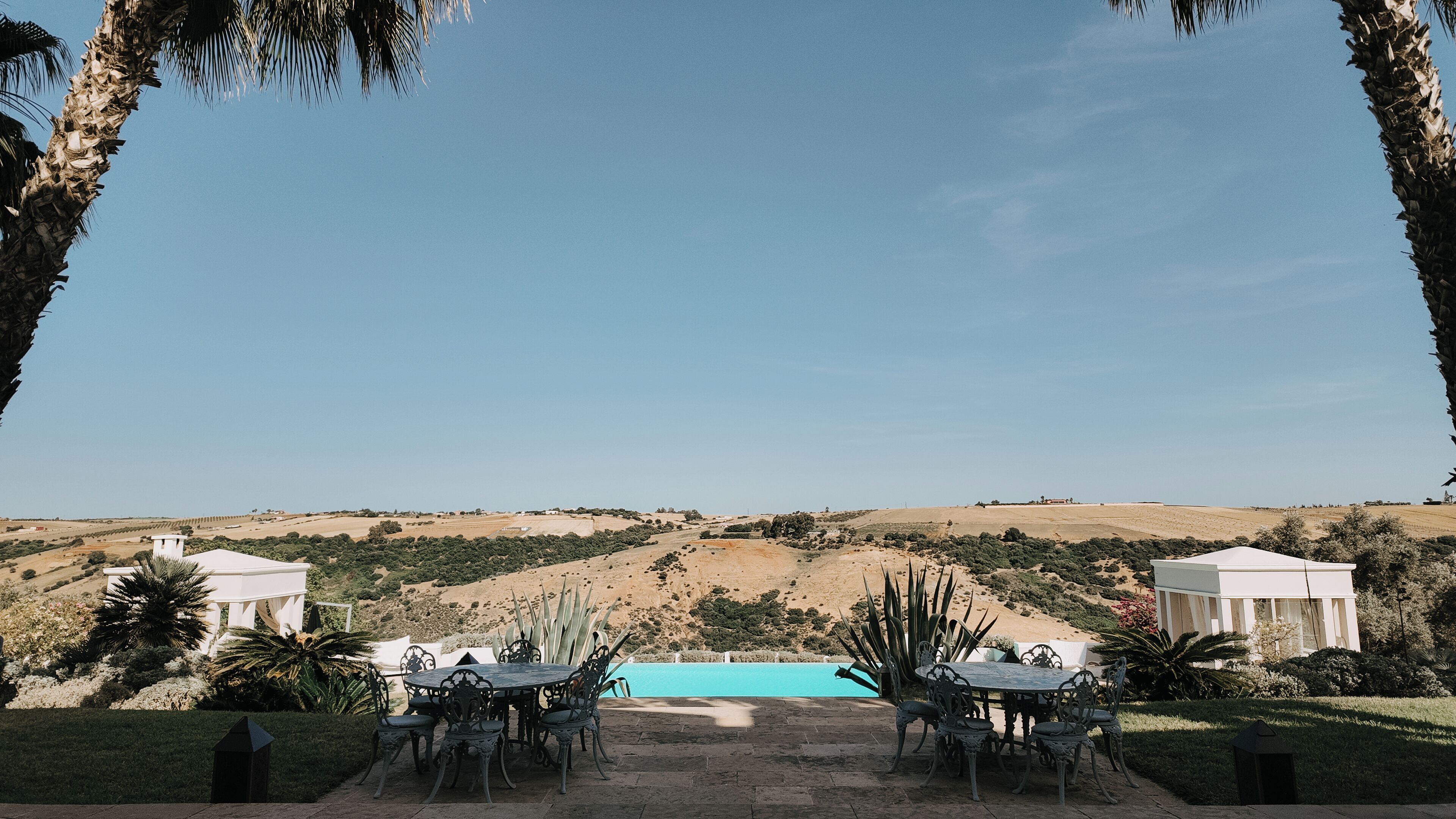 A patio with a pool, surrounded by palm trees and a view of a hilly landscape.