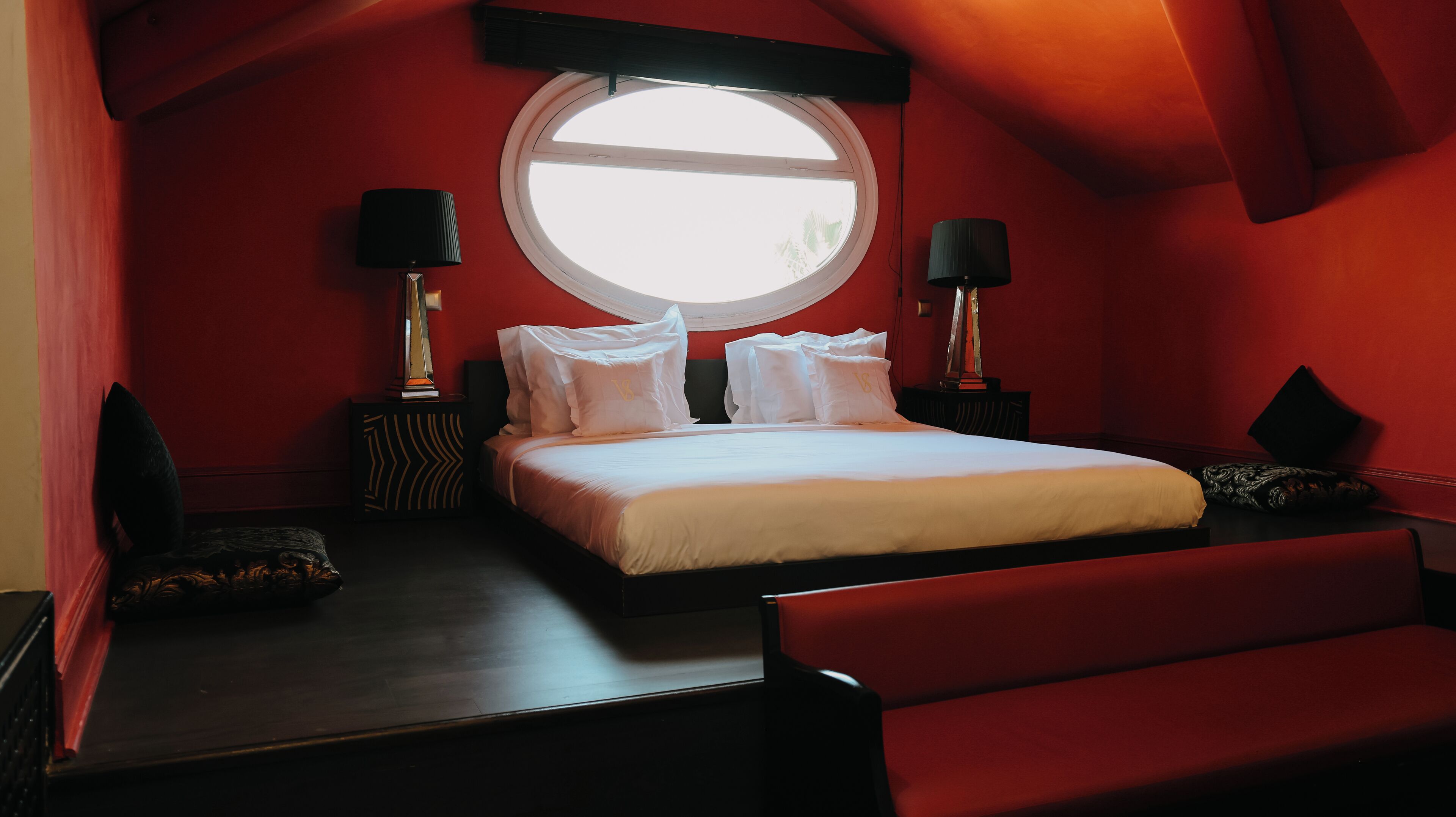 A bedroom with a large round window, a bed with white bedding, two bedside tables with lamps, and a red wall.