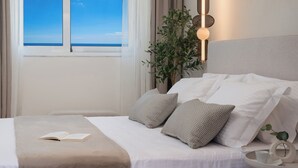 A neatly made bed with white linens and two patterned pillows, a bedside table with a book and a cup, and a view of the sea through a window. - Evilion Home 2 (Agios Nikolaos)