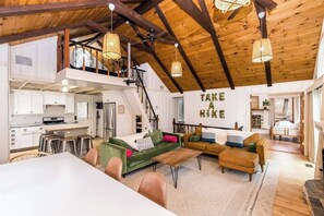 A spacious loft with a high ceiling, exposed wooden beams, a kitchen with white cabinets, a dining area with a table and chairs, and a living room with sofas and a fireplace.