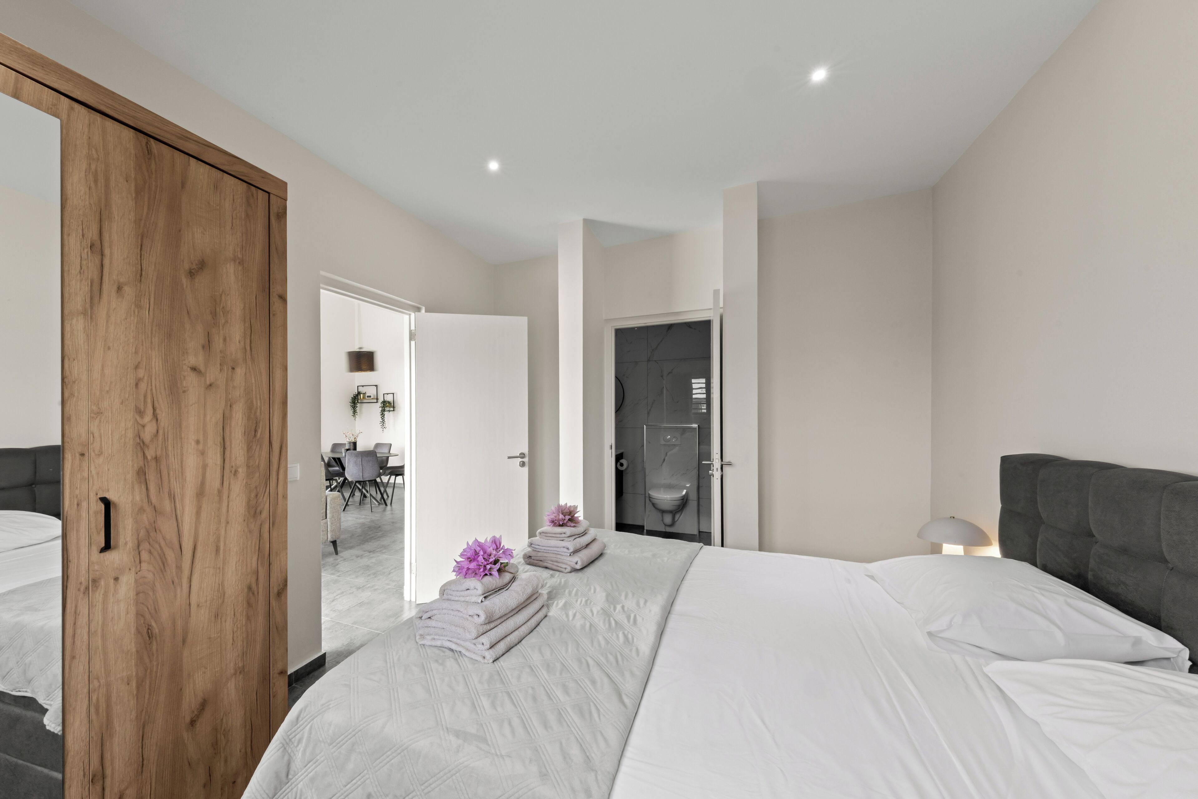 A modern bedroom with a large bed, a wooden wardrobe, a bathroom with a shower, and a dining area visible through an open door.