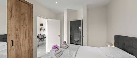 A modern bedroom with a large bed, a wooden wardrobe, a bathroom with a shower, and a dining area visible through an open door.