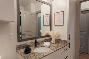 A bathroom with a granite countertop, a white sink, a black faucet, a framed poster, and a mirror. - Walking Paradise-Rooftop Pool-Free Parking & Wifi (Houston)