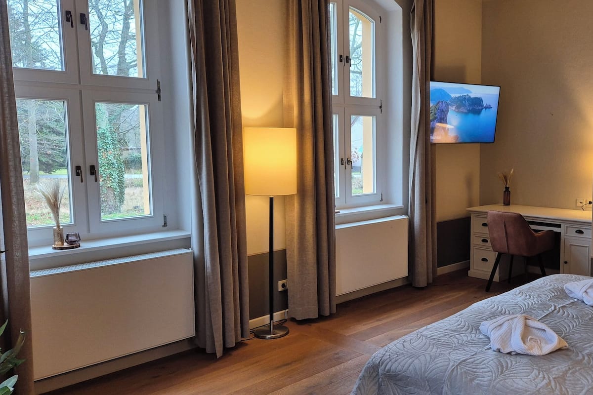 A hotel room with a bed, a desk, a chair, a TV, and large windows.