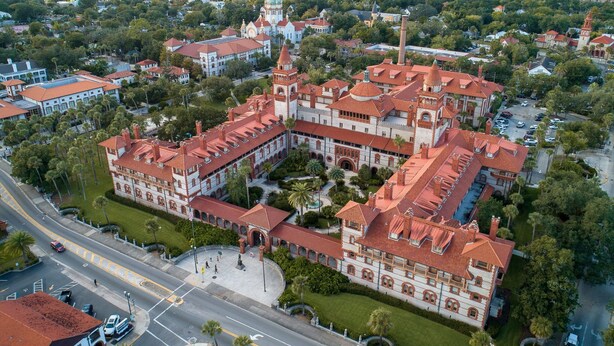 An aerial view of a large, historic building with a central courtyard, surrounded by lush greenery and a river in the background.