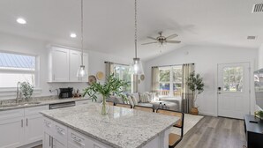A modern kitchen with white cabinets, granite countertops, and a central island. There is a ceiling fan, pendant lights, and a view of the outdoors through a large window.