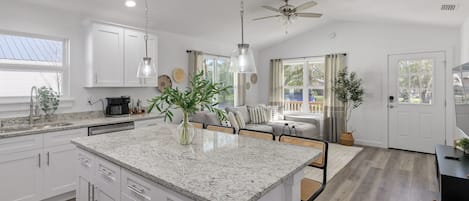 A modern kitchen with white cabinets, granite countertops, and a central island. There is a ceiling fan, pendant lights, and a view of the outdoors through a large window.