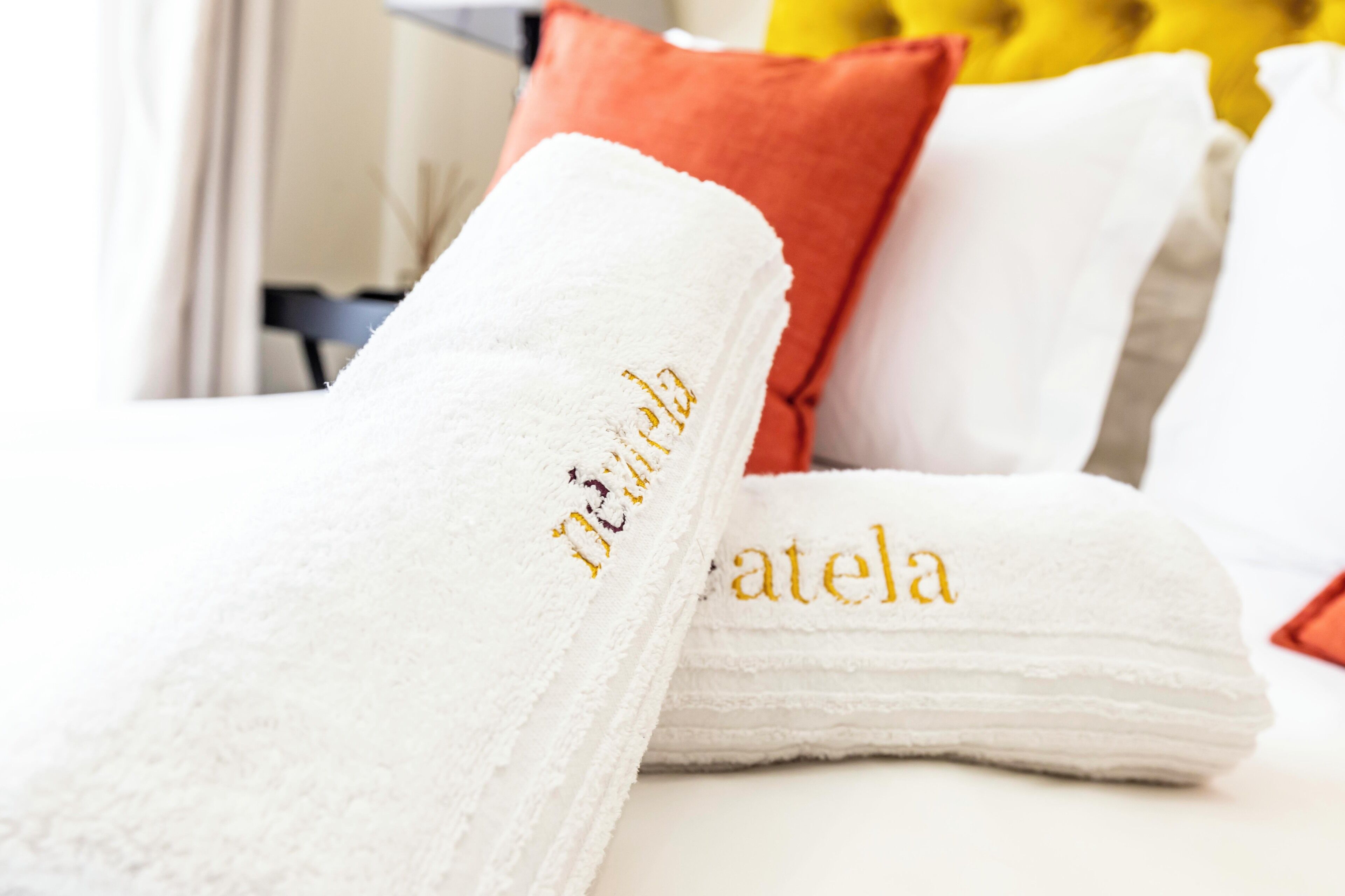 A neatly folded white towel with embroidered text, placed on a bed with a red pillow and a white pillow.
