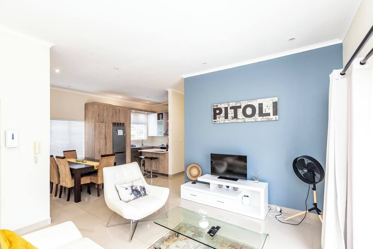 Una sala de estar moderna con una mesa de centro de vidrio, un sofá blanco, un televisor de pantalla plana y un ventilador de pared.