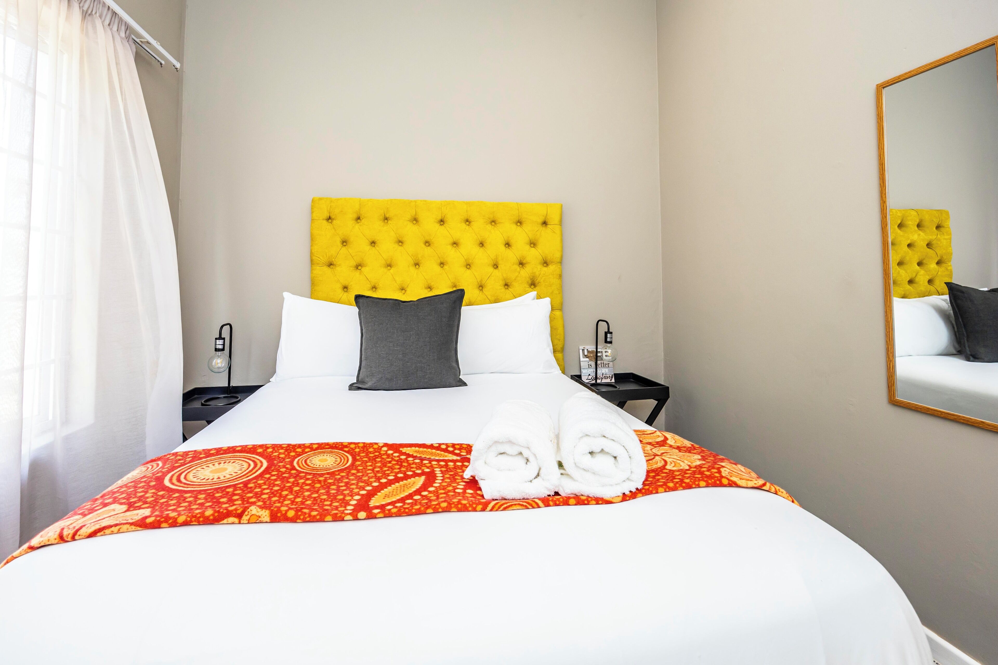 Une chambre d’hôtel avec un grand lit, une tête de lit jaune à motifs capitonnés, du linge de lit blanc, une couvre-lit rouge à motifs et un miroir au mur.