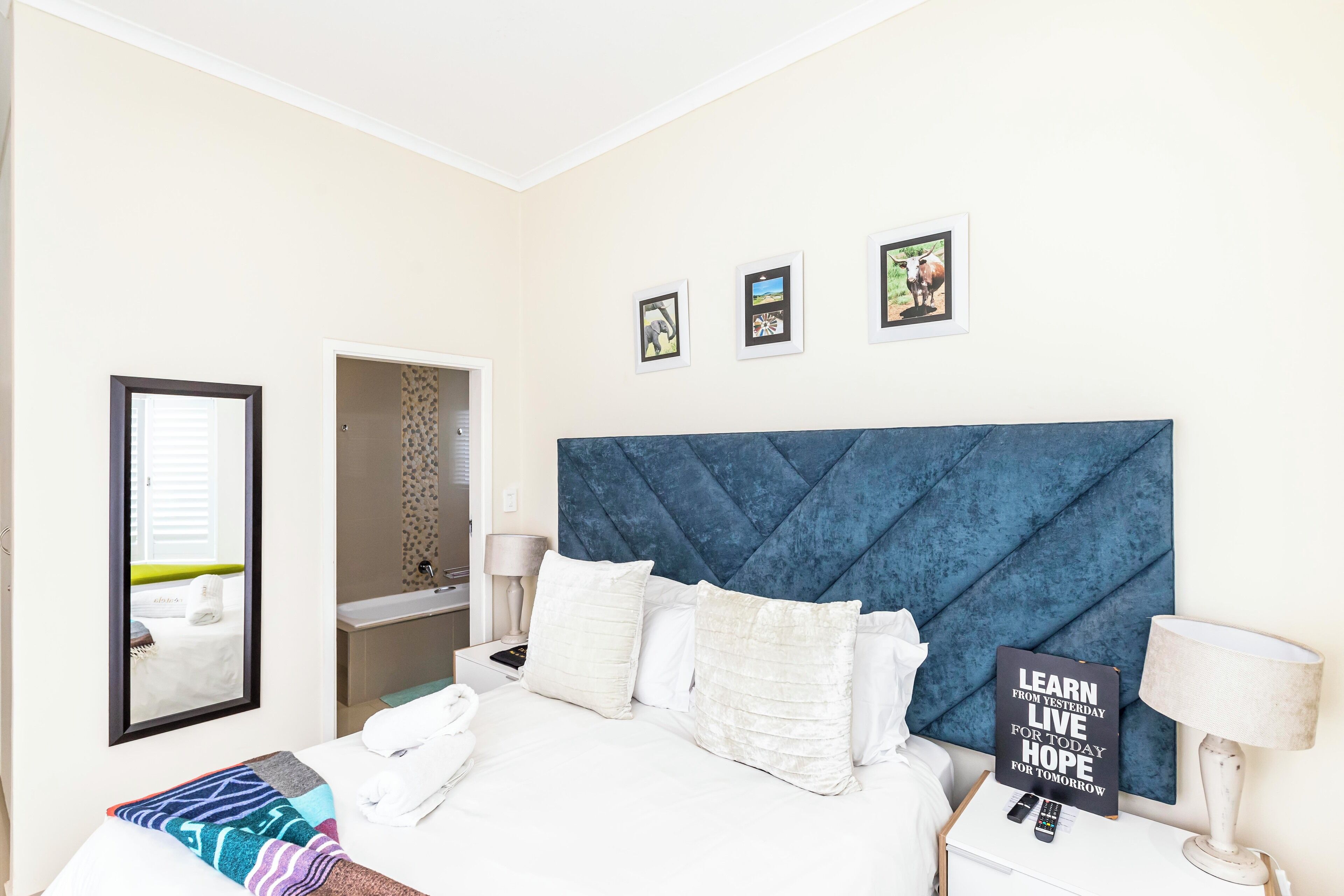 Habitación de hotel con una cama grande, cabecero, mesita de noche con lámpara, espejo y una pared con cuadros enmarcados.